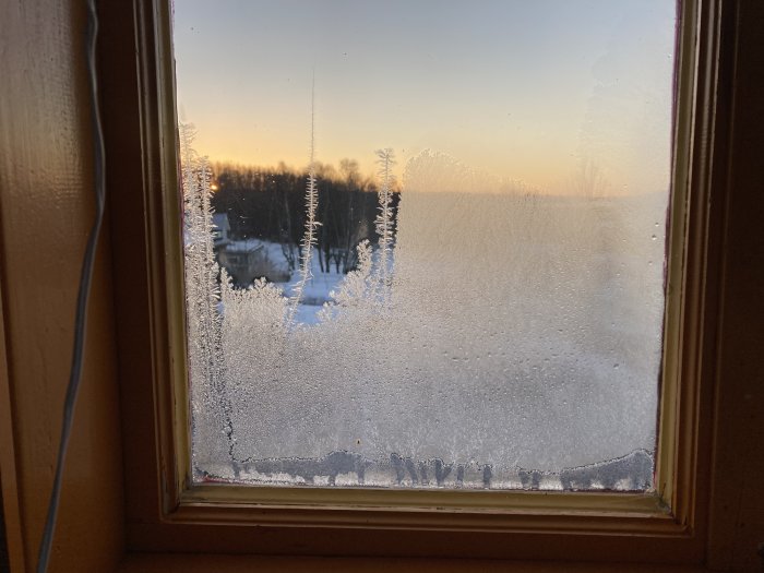 Frostiga kristaller på fönster med vinterlandskap i bakgrunden vid solnedgång.