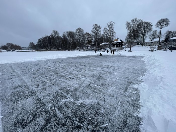 Personer på isbelagd sjö, omgivna av vinterlandskap med snö och träd.