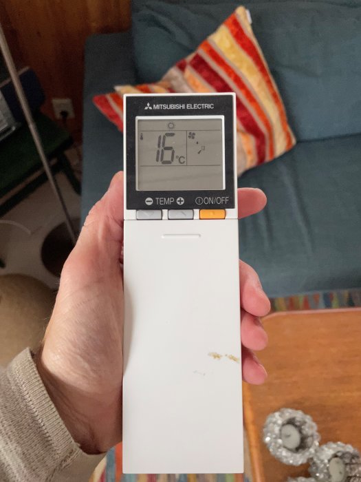 Hand håller Mitsubishi Electric fjärrkontroll, visar 16 grader Celsius, inomhusmiljö, soffhörn och dekorativa kuddar.
