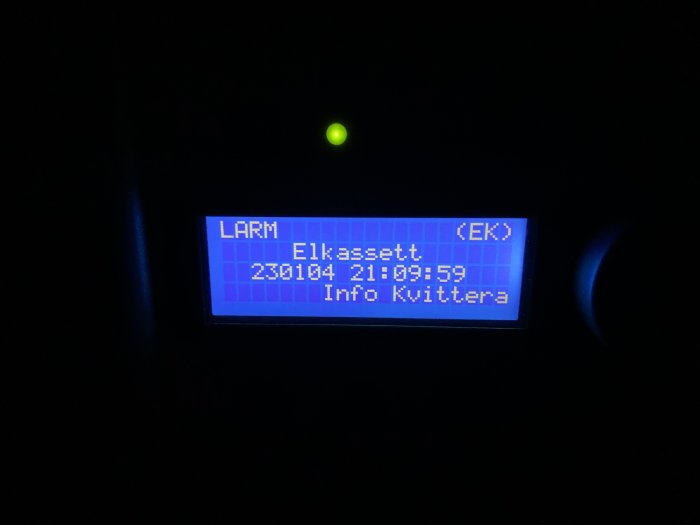 Digital skärm visar larmtext: "Elkassett.", omgiven av mörker. Grünt diodljus synligt ovanför.