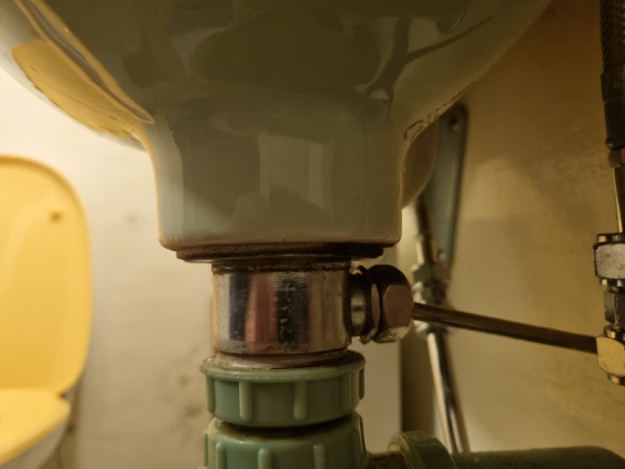 Närbild på en handfatssifon och rörledningar, med gulaktig bakgrund och antydan till toalettstol.