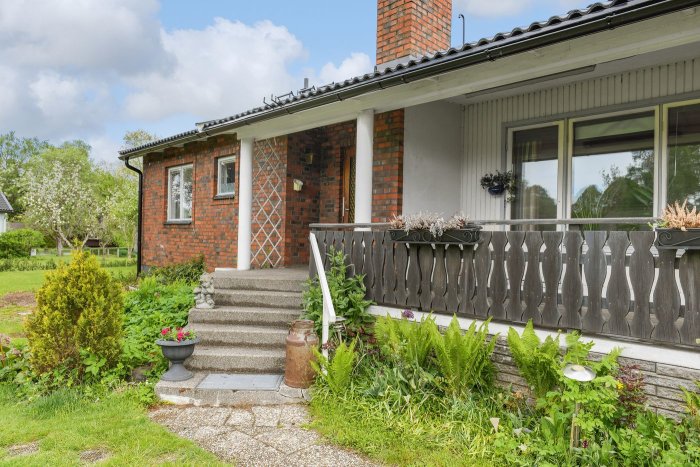 Ett tegelhus med veranda, grönska, trädgård och molnig himmel. Trappa upp till entrén.