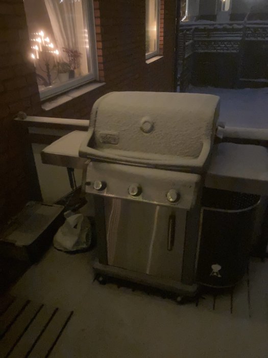 En grill täckt av snö på en balkong vid kvällstid med tända ljus i fönstret.