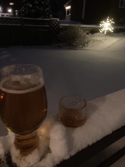 Ölglas på snötäckt räcke, kväll, julbelysning i bakgrunden, lugnt vintermotiv.