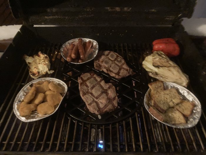 Grill med kött, korv, grönsaker och folieformar med tillbehör. Utomhus, mörkt, verkar vara kallt ute.