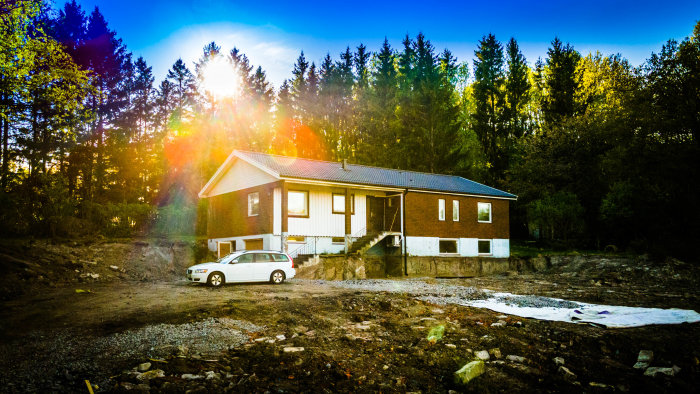Ett hus och en bil framför, omgivna av skog, under en dramatisk solnedgång med lens flare.