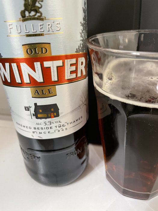 Fuller's Old Winter Ale-flaska nära halvfullt ölglas på ljust underlag.