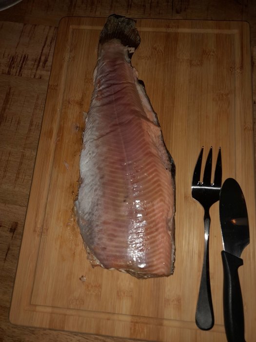 Ett stort stycke fiskfilé på en skärbräda med gaffel och kniv bredvid.