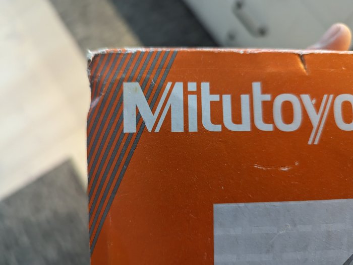 Orangefärgad förpackning med texten "Mitutoyo" i förgrunden, hållen av person, oskarp bakgrund.