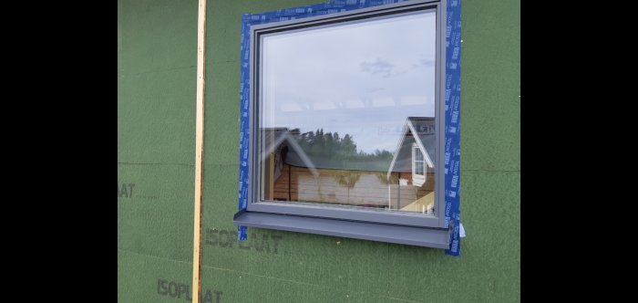 Ett nyinstallerat fönster med isoleringstejp på en grönbeklädd väggyta, träribba till vänster, reflektion av himmel.