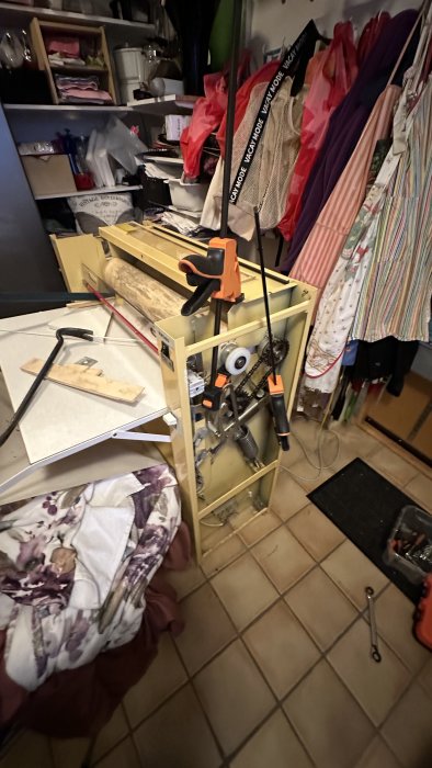 Oorganiserad verkstad med verktyg, träbitar och tyger. Verkar vara i en källare eller garage.