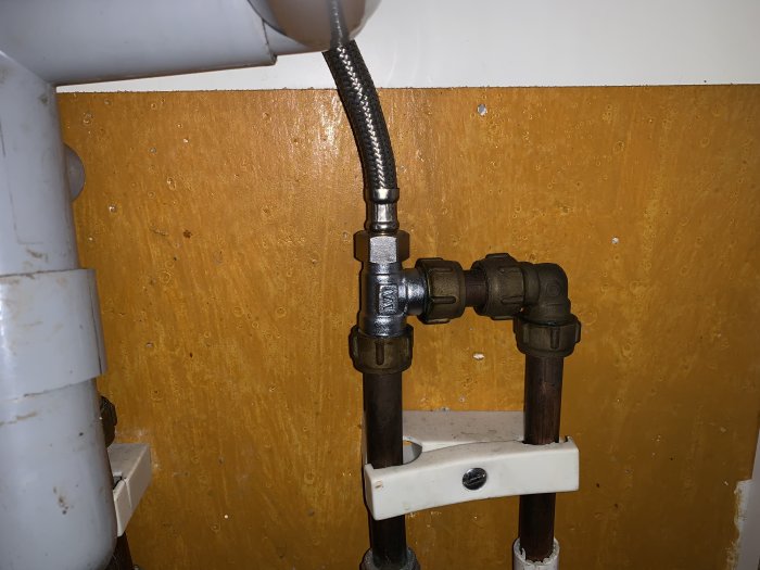 VVS-installation med rör, ventiler och anslutningsslang under en diskbänk mot en trävägg.