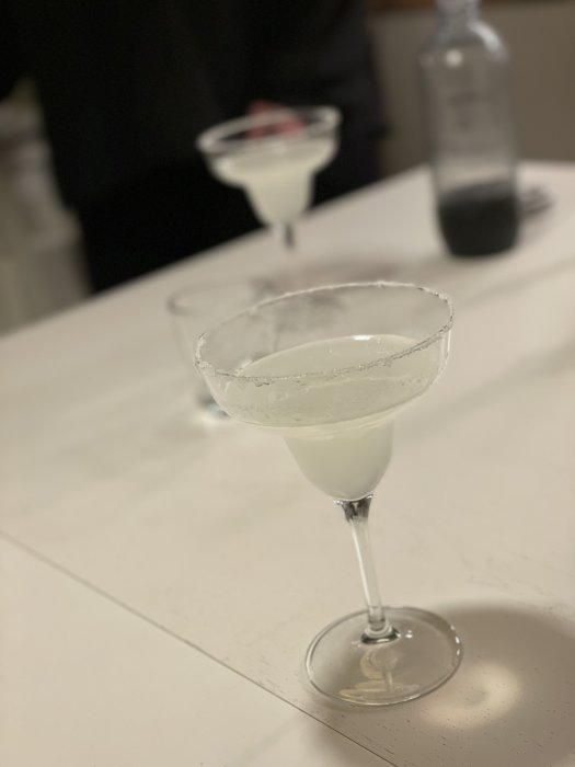 Två margarita-cocktails med saltade kanter på barmiljö, flaska i bakgrunden, fokus på främre glaset.