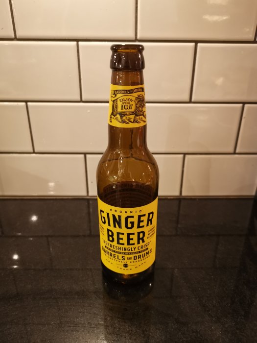 En brun glasflaska märkt "GINGER BEER" på ett grått köksbord med vita kakelplattor i bakgrunden.