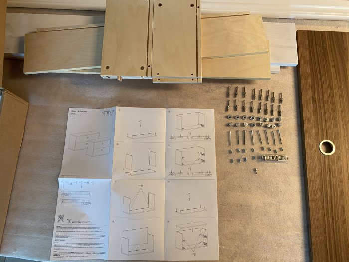 Instruktionsblad och omonterade delar av en byrå på ett golv, redo för ihopsättning, med skruvar och fästelement.