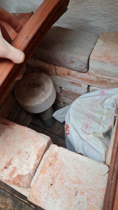 Hand lyfter tegel, visar rörsystem och påse under golvet. Renovering eller underhåll i byggnad verkar pågå.