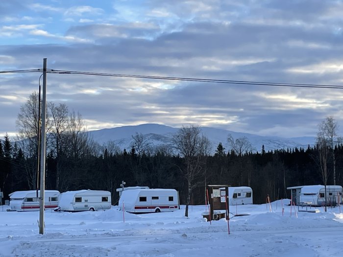 Campingplats med husvagnar i snö, berg i bakgrunden, träden, molnig himmel vid skymning eller gryning, elledningar.