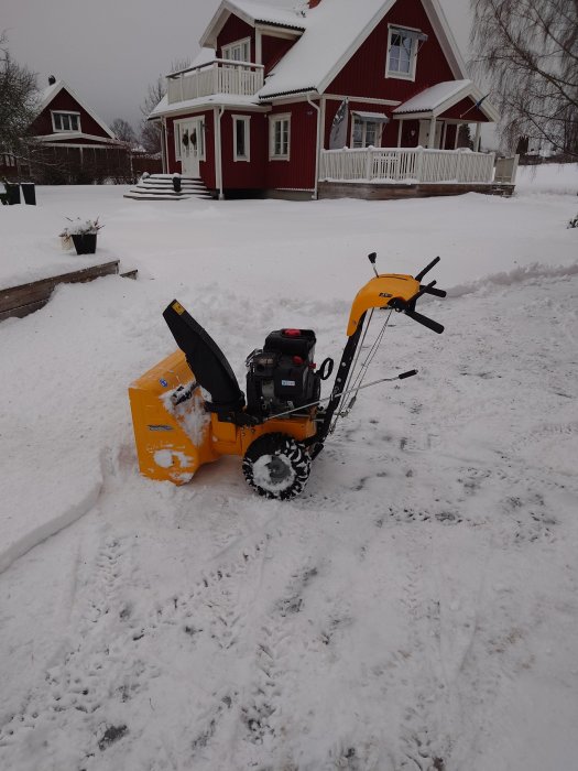 Snöslunga framför rött hus med vit snö, grå himmel, vinterdag, trädgård, trappsteg täckta av snö.