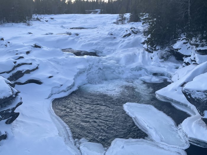 Vinterlandskap med delvis frusen flod, snötäckta klippor och skog i bakgrunden.