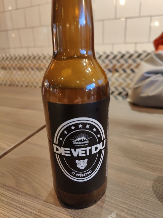 Brun glasflaska med etikett "DEVETDU BY BRÖDERNAS" på köksbänk, kakel i bakgrunden.