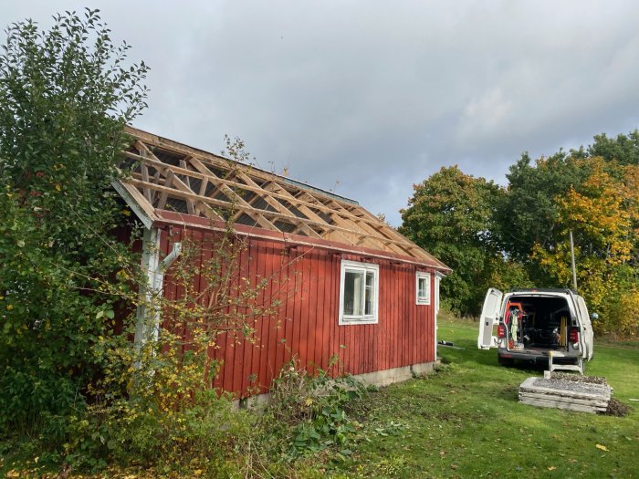 Rött hus under renovering, delvis saknar tak, öppen skåpbil, höstigt landskap, verktyg, grönska.
