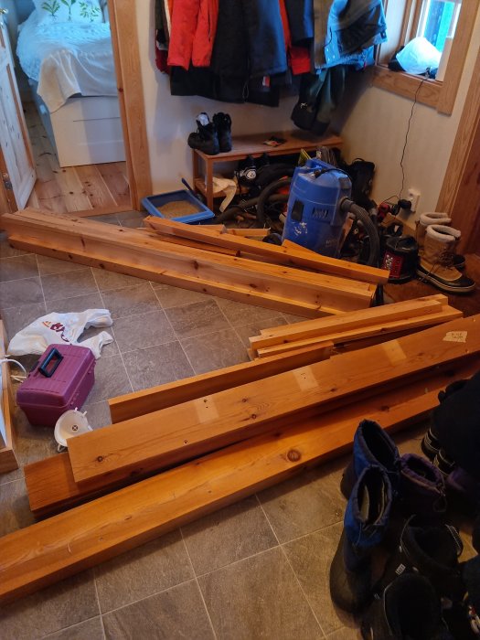 Träplankor på golv, uppställda möbler, jackor, skor och dammsugare i rum, renoveringsprojekt.