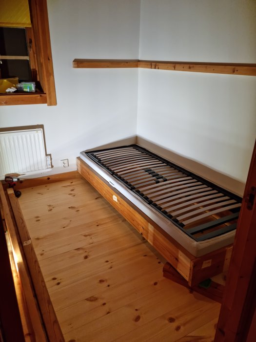 Ett sovrumshörn med tom sängstomme och ribbotten, trägolv, vit vägg, och radiator. Inga sängkläder eller madrass synliga.