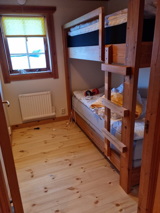 Trävåningssäng i litet rum, fönster, trägolv, huvudtelefoner på säng, osorterade föremål, mysigt inredd.