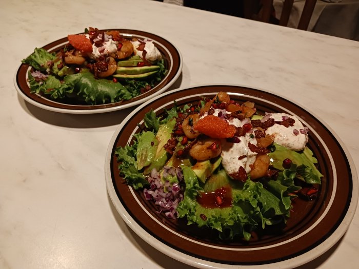 Två salladsrätter med gröna blad, avokado, rödbetor, dressing och strössel, serverade på marmor bord.