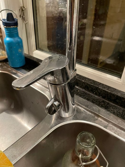 Kökskran över diskho, fönster, blå vattenflaska, diskmedelsflaska, viss reflexion och belysning.