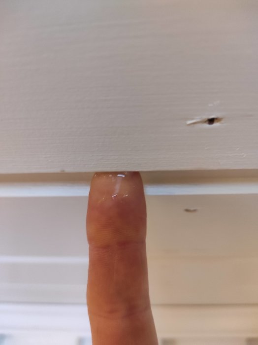 Ett finger pekar på en vitmålad yta med en liten skada eller spricka.
