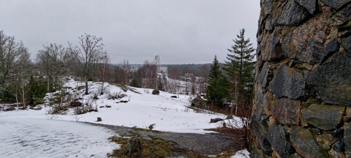 Vinterlandskap med snötäckt mark, kala träd, stenmur till höger och en byggnad i bakgrunden.