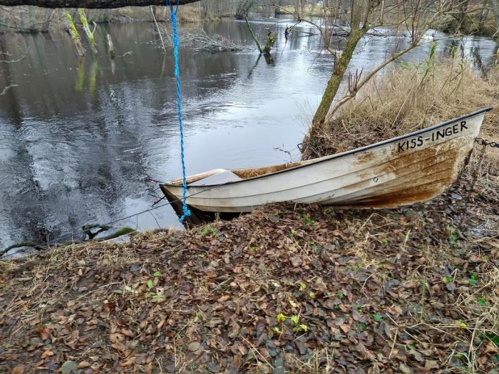 En förtöjd roddbåt vid en flodkant omgiven av löv och träd, spegling i vattnet.