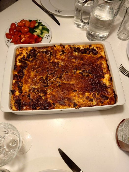 Gratinerad lasagne i form, salladstallrik, vattenglas, vitt matbord, informell måltidsmiljö.