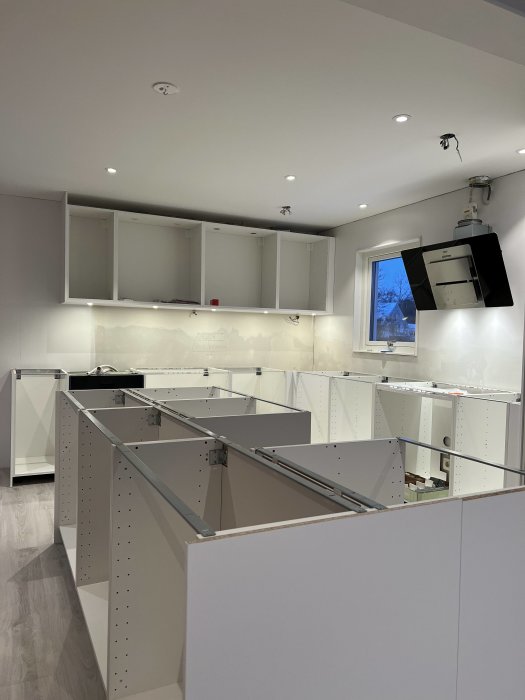 Ett modernt kök under installation med vita skåp och tomt golv, under takbelysning och dagsljus från fönstret.