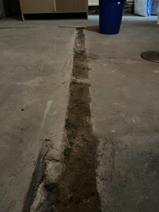 En grå betonggolv med en utgrävd ränna, smuts och en blå behållare i bakgrunden.
