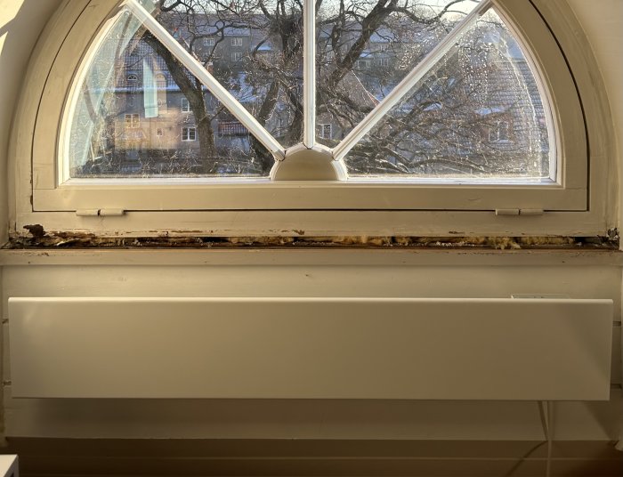 Bågformat fönster med utsikt, skadad fönsterkarm, solsken, inomhus, radiator under fönster, träd och byggnader utanför.
