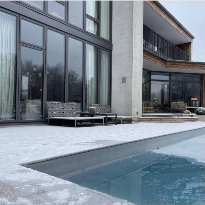 Modern husfasad med stora fönster, terrass, pool, snö och utemöbler.