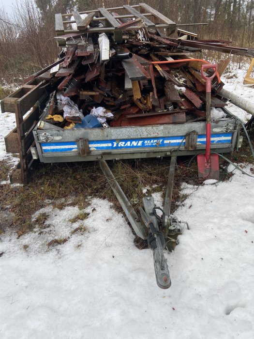 Släpvagn överfylld med skrotat material och byggavfall, står på snötäckt mark.