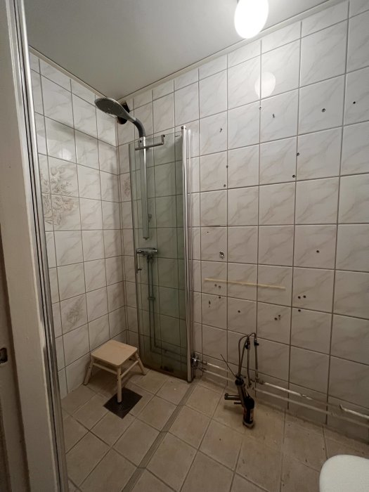 Ett badrum med duschhörna, vita kakelväggar, träpall och golvstående blandare.
