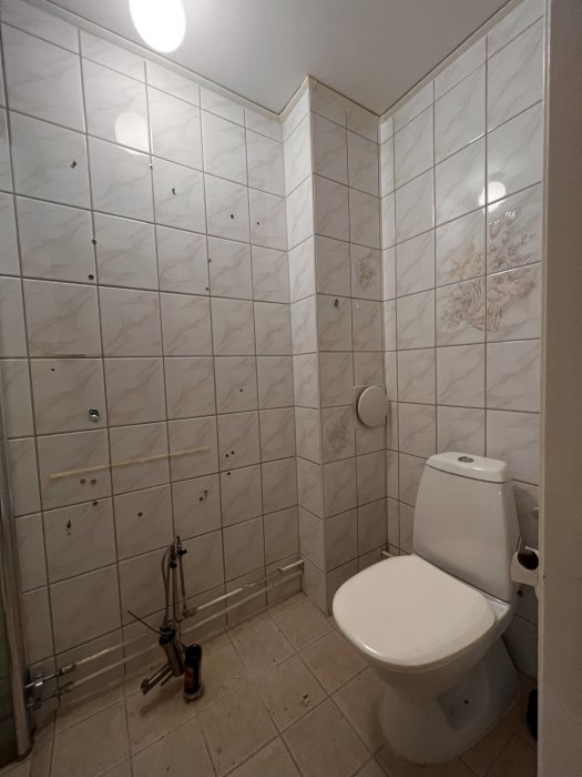Ett badrum med toalett under renovering, saknar handfat, kakelväggar, verktyg på golvet.