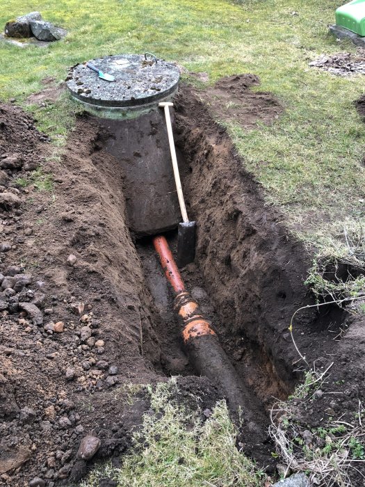 Grävd ränna i gräsmatta med synlig orange rörledning och spade mot brunnslock.