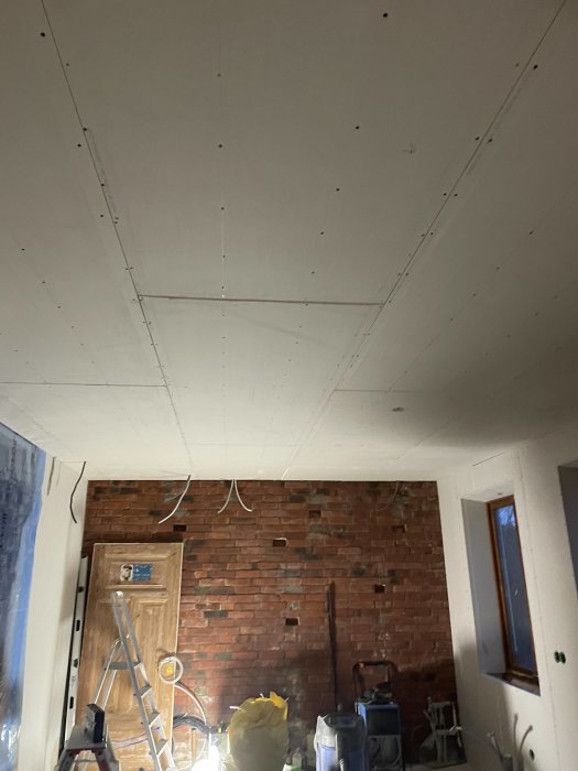 Inomhusrenovering pågår, gipsskivor i tak, tegelvägg, stege och byggmaterial, ospacklade skruvhål synliga.