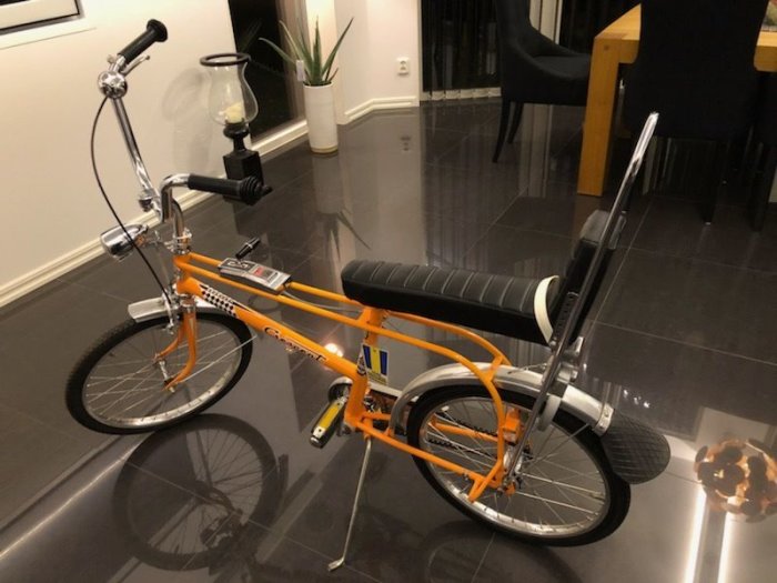 Orange choppercykel i inomhusmiljö med högt styre och lång sadel.