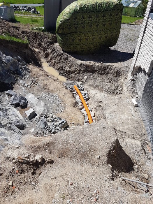 Grävning nära byggnad, orangefärgade rör syns, vattenansamling, ojämn terräng, sten och grus, solljus, grönt gräs i bakgrunden.