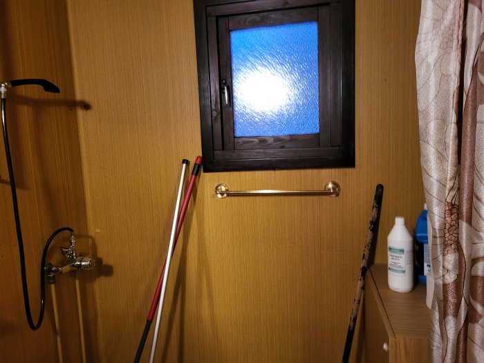 Ett duschutrymme med greppvänliga stänger och ett färgat fönster. Städredskap och rengöringsmedel är synliga.