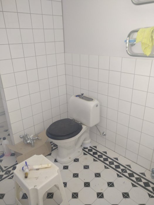 En toalett med vit och svart kakel, säkerhetshandtag, pall och renoveringsmaterial.