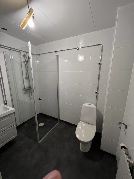 Ett modernt badrum med duschhörna, toalett, vita kakelväggar och mörkt golv.