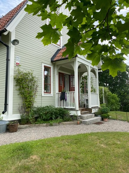 Grönt hus med vit veranda, röd dörr, krukväxter, grusgång, lummiga trädgrenar i förgrunden.