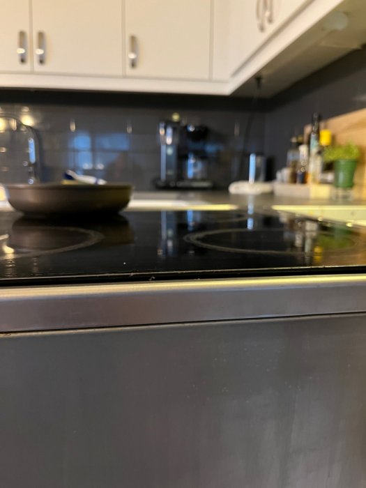 Ett kök med spishäll, stekpanna, skåp, espressomaskin och kryddväxter. Oskärpa i förgrunden, detaljer i bakgrunden.
